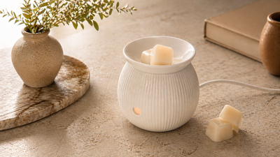 White ceramic wax warmer with melts on a stone surface, next to a vase with greenery and a book.